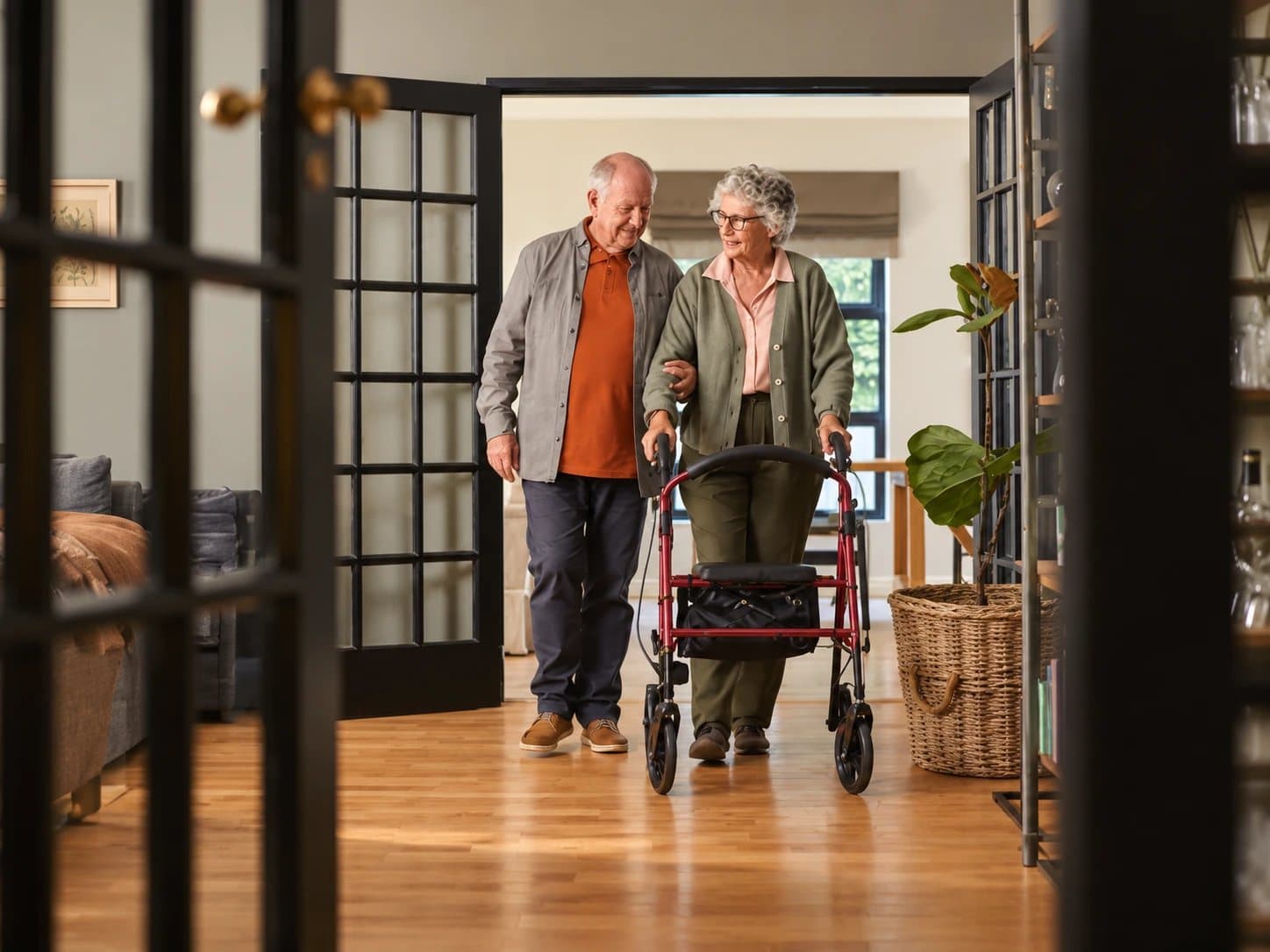 An older couple walking together through a hallway at home, one of them using a walking frame