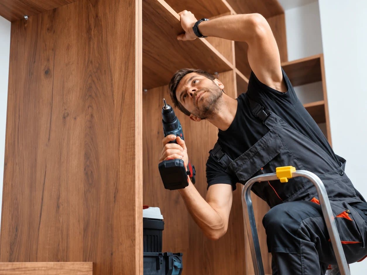 A tradesperson on a stepladder fitting a shelf in a built-in timber wardrobe with a cordless drill